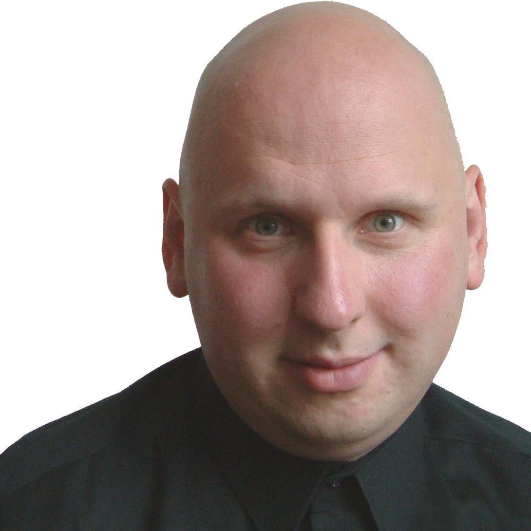 Ein Mann mit Glatze und heller Haut, der ein schwarzes Hemd trägt und mit neutralem Blick direkt in die Kamera schaut.