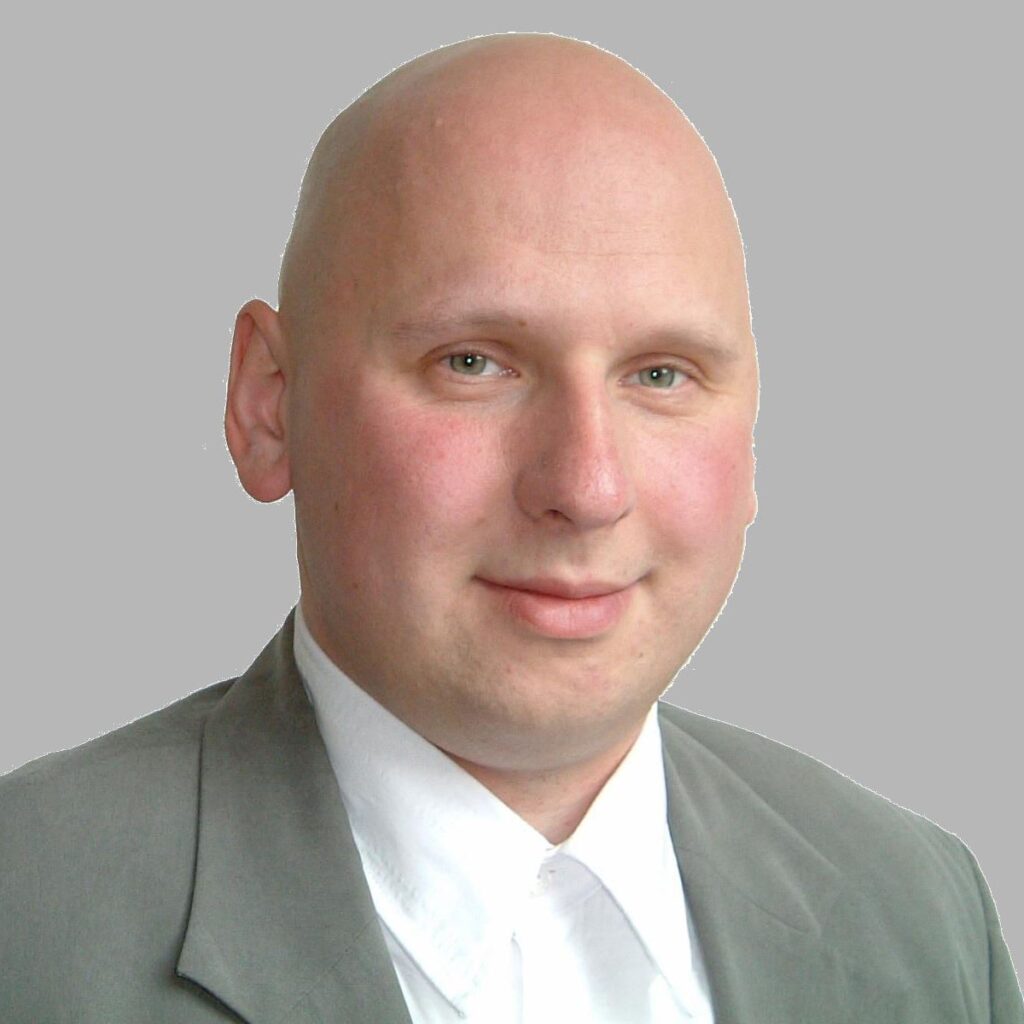 Glatzköpfiger Mann in grauem Anzug und weißem Hemd, lächelnd, vor neutralem grauem Hintergrund.